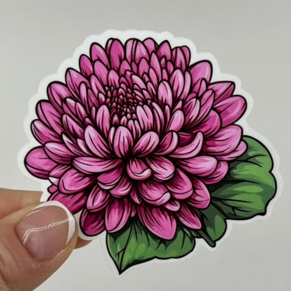 Fingers holding a sticker of a pink aster with green leaves.