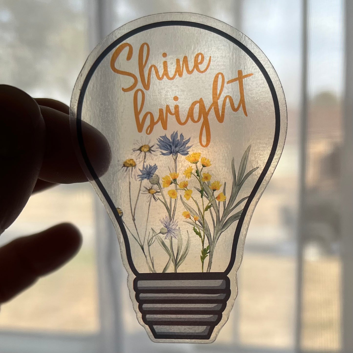 A clear vinyl sticker with a design of a lightbulb and flowers, and the text 'Shine bright'.