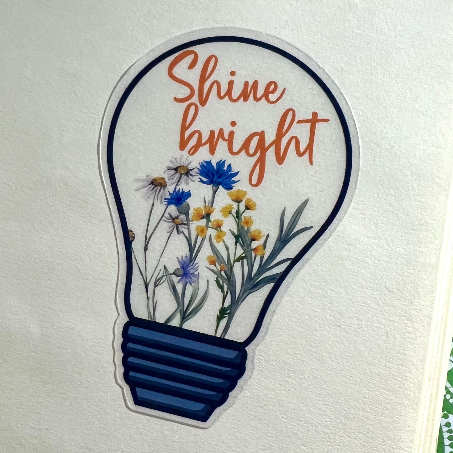 A clear vinyl sticker with a design of a lightbulb and flowers, and the text 'Shine bright'.
