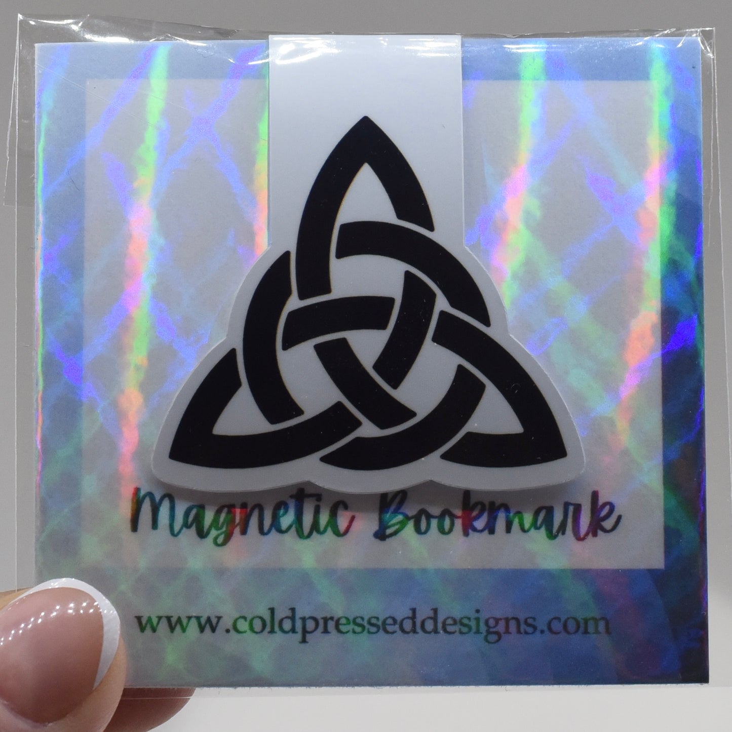 A hand holding a glossy, double-sided magnetic bookmark with a triquetra symbol design.