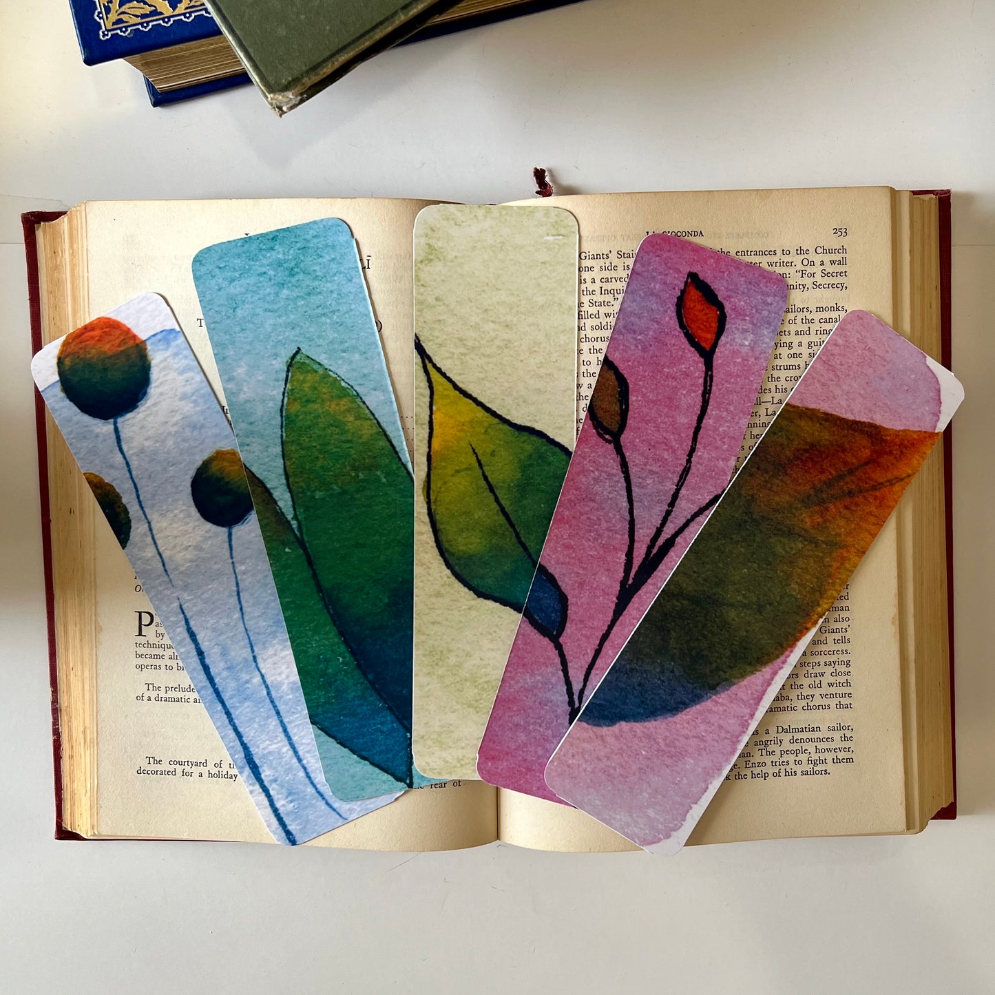 A set of five glossy handmade bookmarks with watercolor floral doodles in various colors placed on an open book.