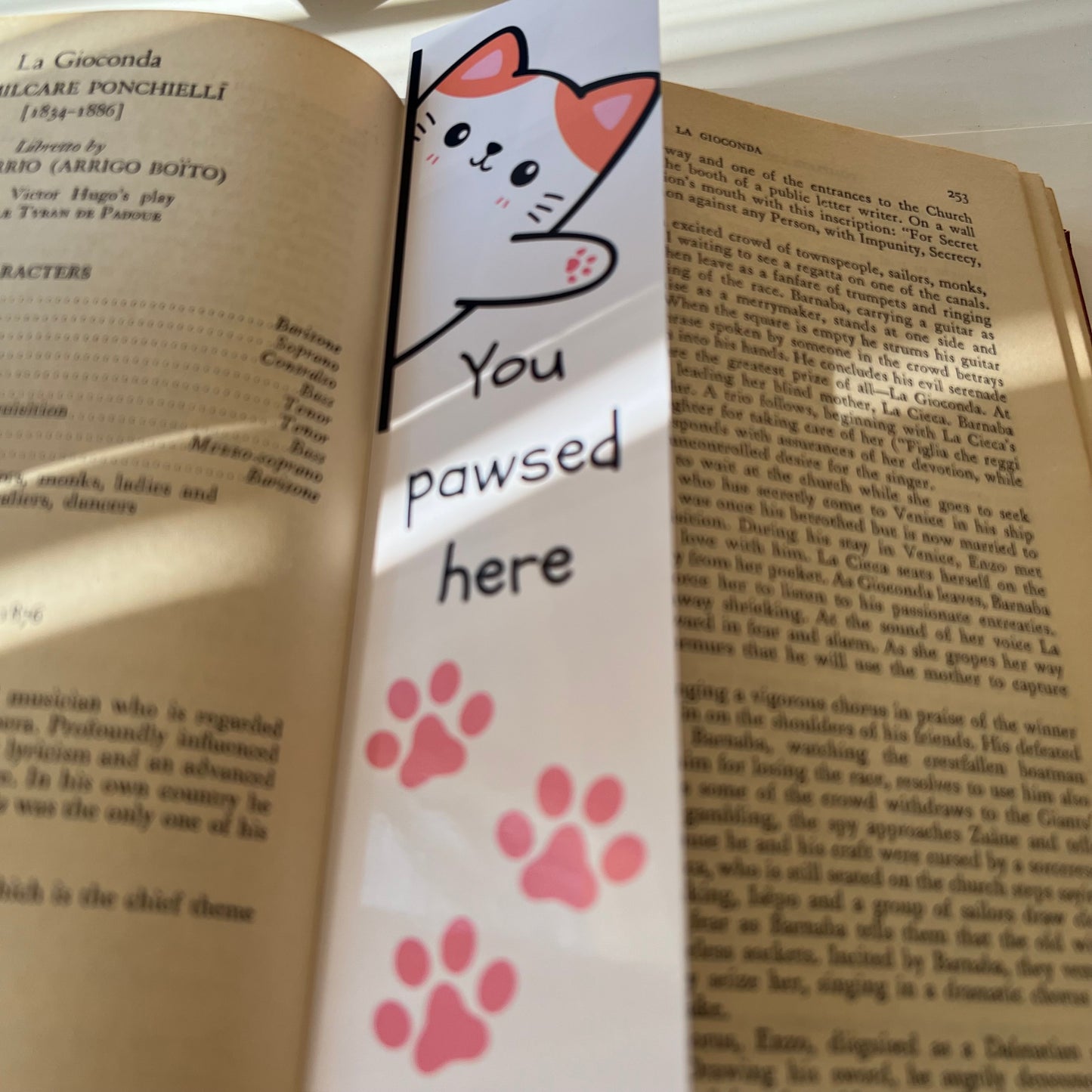 A rectangular bookmark with a cartoon cat face, the phrase 'YOU PAWSED HERE' and pink paw prints on a book.