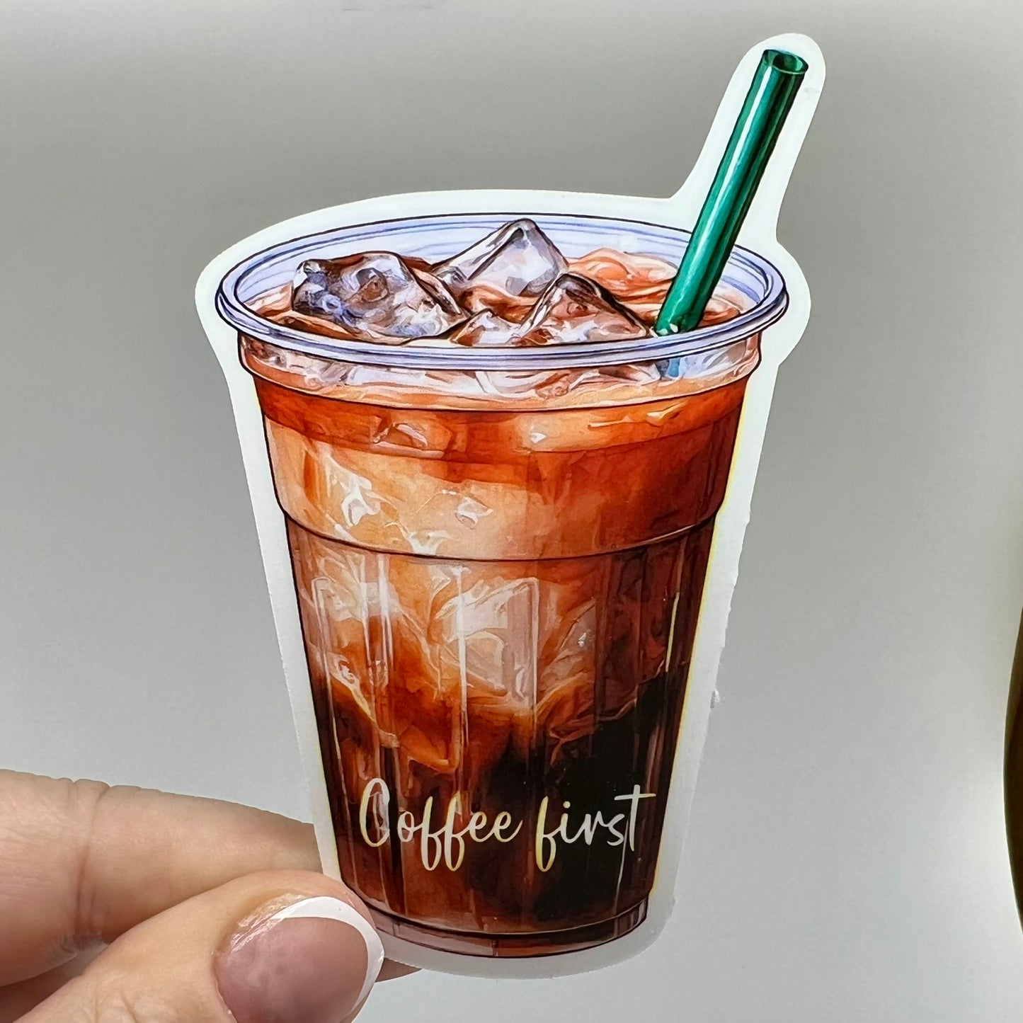 A vinyl sticker of an iced coffee cup with 'coffee first' written on it. The sticker is brown with a clear cup, green straw, and coffee and milk colored beverage inside.