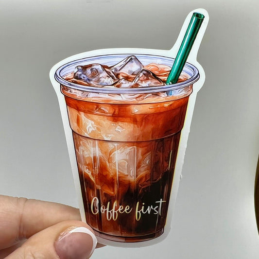 A vinyl sticker of an iced coffee cup with 'coffee first' written on it. The sticker is brown with a clear cup, green straw, and coffee and milk colored beverage inside.