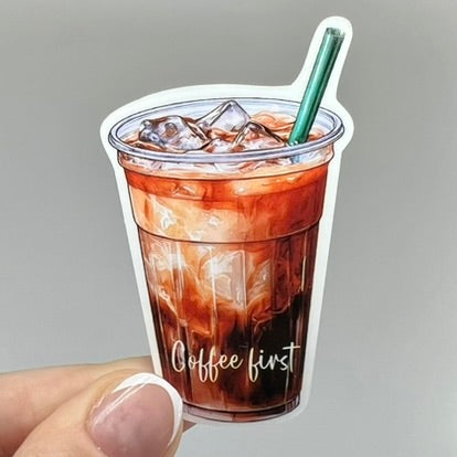 A vinyl sticker of an iced coffee cup with 'coffee first' written on it. The sticker is brown with a clear cup, green straw, and coffee and milk colored beverage inside.