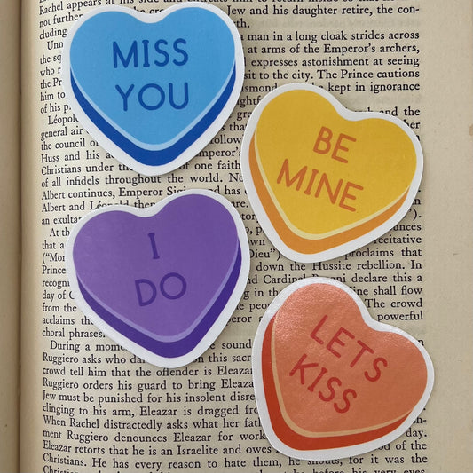 A set of four vinyl stickers with heart-shaped designs and various affectionate phrases written on them, placed on a book page.