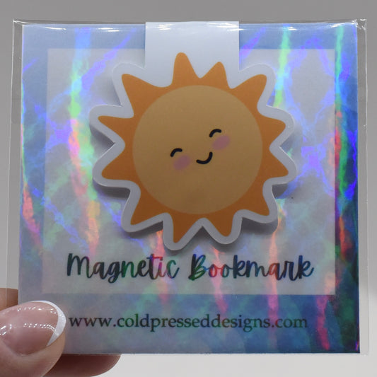 A hand holding a glossy, double-sided magnetic bookmark in a yellow sun design with a kawaii face and the text 'Magnetic Bookmark' on a reflective background.
