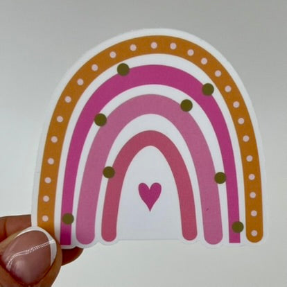 A vinyl sticker with an orange and pink rainbow design and small heart shapes; held by a hand against a white background.