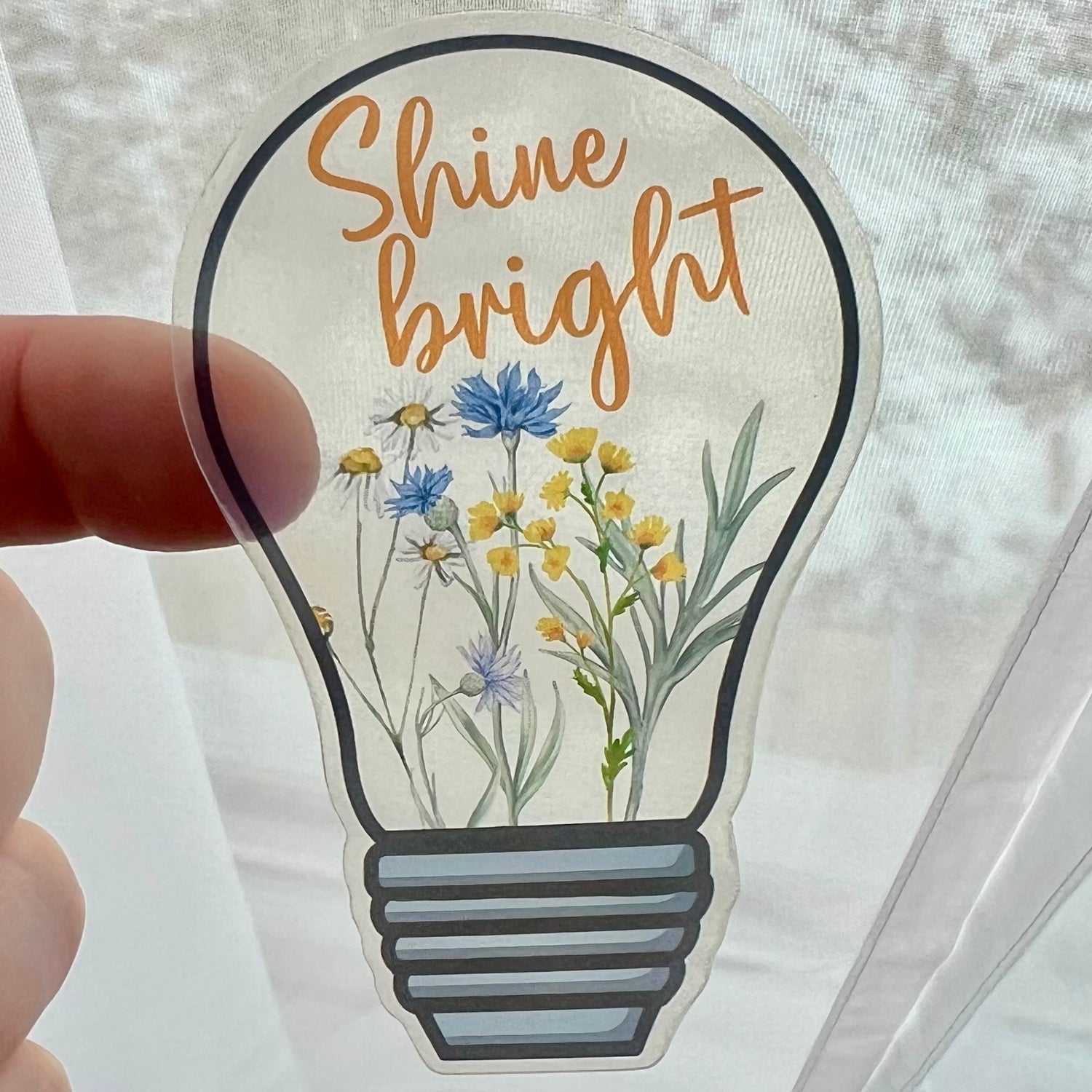 A clear vinyl sticker with a design of a lightbulb and flowers, and the text 'Shine bright'.