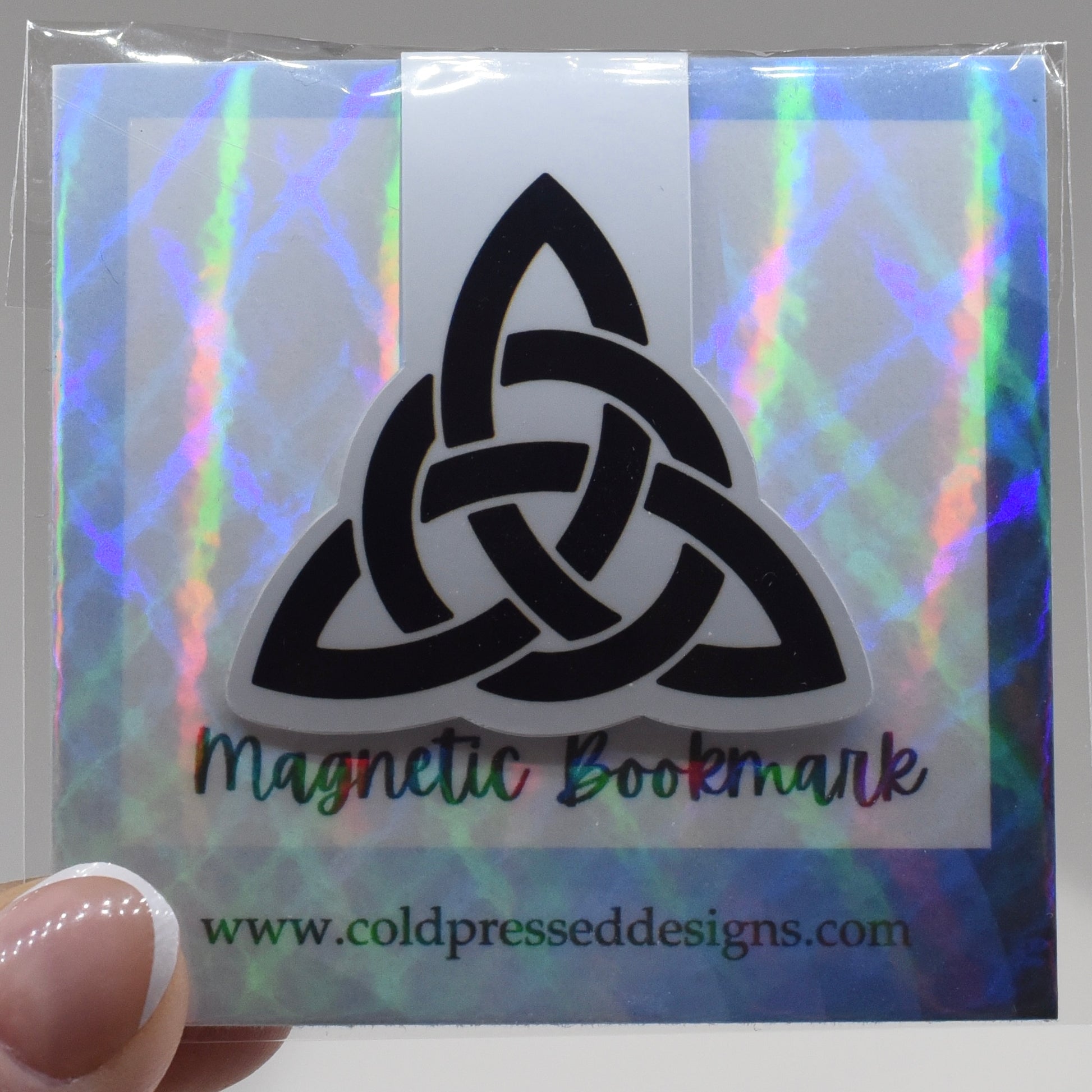 A hand holding a glossy, double-sided magnetic bookmark with a triquetra symbol design.