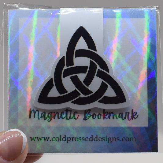 A hand holding a glossy, double-sided magnetic bookmark with a triquetra symbol design.