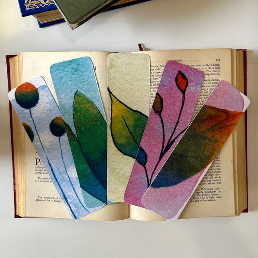 A set of five glossy handmade bookmarks with watercolor floral doodles in various colors placed on an open book.